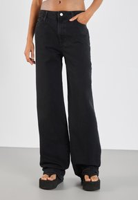 Personne portant un pantalon noir taille haute à jambes larges, une main dans la poche et pieds nus sur des sandales à plateformes noires.