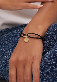 Braccialetto con cordoncino nero, chiusura in metallo dorato e ciondolo con logo inciso. Il cordoncino è testurizzato e regolabile.