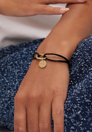 Braccialetto con cordoncino nero, chiusura in metallo dorato e ciondolo con logo inciso. Il cordoncino è testurizzato e regolabile.