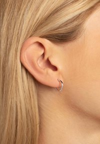 Piccolo orecchino a cerchio d'argento, forma rotonda, texture liscia, esposto sull'orecchio di una persona, con capelli biondi lisci sullo sfondo.