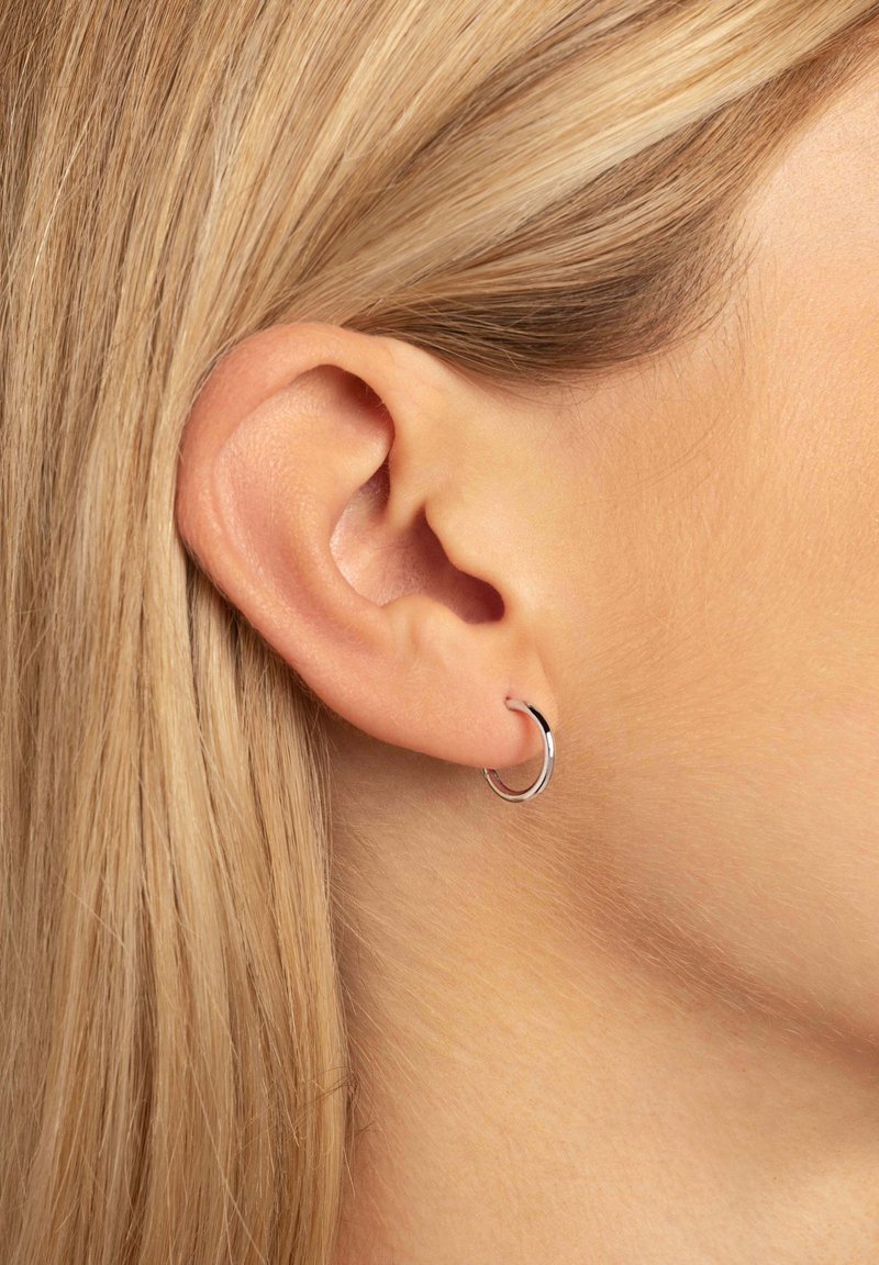 Piccolo orecchino a cerchio d'argento, forma rotonda, texture liscia, esposto sull'orecchio di una persona, con capelli biondi lisci sullo sfondo.