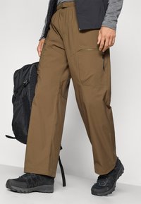 Pantalons d'extérieur légers de couleur marron avec une coupe décontractée, une taille élastique, des poches latérales et des accents zippés subtils. Associés à des chaussures noires et un sac à dos.