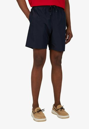 Homme portant un short bleu marine à cordon, une chemise rouge et des chaussures décontractées beiges avec des lacets noirs, debout les mains dans les poches.