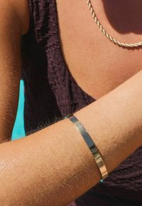 Bracelet jonc en or et argent sur un bras bronzé, présentant un design lisse et poli avec un profil plat et un éclat métallique.