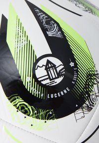 Balts futbols ar melniem un neona zaļiem grafiskajiem rakstiem, ar apļa logotipu, kurā attēlots būves dizains un koordinātas.