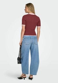 Femme de dos portant un haut ajusté à manches courtes couleur bordeaux, un jean bleu à jambes larges, des talons slingback noirs et tenant un sac à main noir.