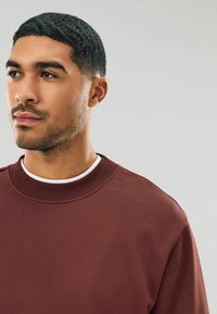 Hanorac maron cu guler crenelat, textură moale și croială lejeră. Purtați peste un tricou alb, are un design minimalist.