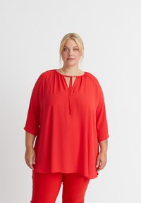 Rote Bluse mit lockerer Passform, rundem Ausschnitt und Dreiviertelärmeln. Hat ein Schlüssellochdetail vorne und eine glatte Stofftextur.