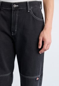 Pantaloni din denim negri cu coasere albe, design clasic cu cinci buzunare și o etichetă cu logo pe coapsa stângă. Materialul pare durabil și are o textură netedă.