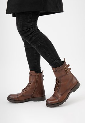 Bruine leren enkelboots met veters en een zijrits, voorzien van twee gespsieraden en een gestructureerde afwerking. Robuuste zwarte rubberen zool.
