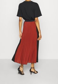Falda larga plisada en rojo profundo y negro, con tejido ligero y un diseño fluido. Combinada con tacones negros con tiras, vista desde la parte posterior.