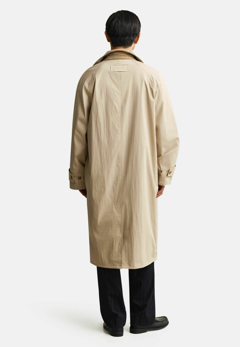 Mann trägt beigefarbenen Trenchcoat, schwarze Hose und schwarze Schuhe, steht mit dem Rücken zu einem einfarbigen weißen Hintergrund.