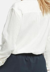 Vit långärmad skjorta i linne, med rygggördel, veck och rullade manschetter, kombinerad med mörka byxor med elastisk midja.