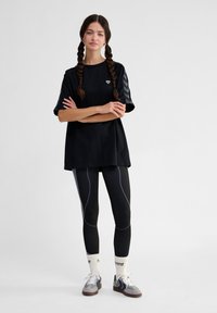 Melnas oversized t-kreklu ar tonāliem chevron piedurknēm, apvienots ar melniem teksturētiem legingiem ar zilu akcentu līnijām un pelēkām sporta apaviem ar tumšiem akcentiem.