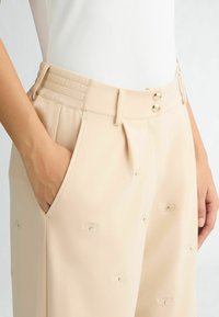 Beige Hose mit hoher Taille, Falten und seitlichen Taschen. Merkmale metallische Knöpfe und ein dezentes eingeprägtes Muster. Glatte Stoffoberfläche.