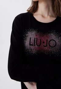 Person trägt ein schwarzes Langarmshirt mit einem gepunkteten Farbverlaufsmuster und dem Text "LIU•JO" auf der Brust, die Arme verschränkt.