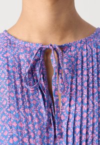 Lilla og lyserødt mønstret stof med folder, der har en bindelukning ved halsudskæringen og en glat tekstur.