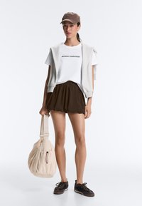 Young woman wearing a "Monday Survivor" white T-shirt, brown shorts, gray sleeveless hoodie, brown cap, black sneakers, holding a beige bag.