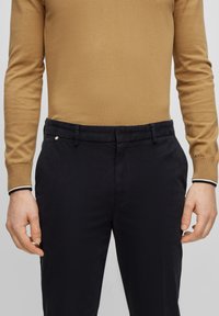 Muž v béžové košili s dlouhým rukávem, zasunuté do černých oblekových kalhot, stojící s rukama u boků.