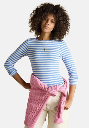 Kobieta o kręconych włosach, ubrana w niebiesko-białą koszulę w paski z długim rękawem, beżowe spodnie oraz różowy sweter zawiązany wokół talii.