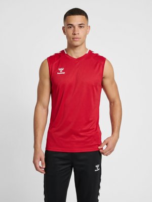 Młody mężczyzna w czerwonej bezrękawnikowej koszulce sportowej i czarnych spodniach sportowych stoi na jednolitym jasnym tle.