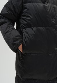 Zwarte bomberjack met een doorgestikt ontwerp, voorzien van een grote voorklep en een zachte, gladde textuur. Inclusief knopenaccenten op de flap.