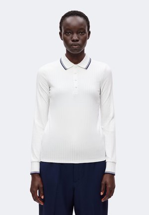 Femme aux cheveux courts portant un polo blanc côtelé à manches longues avec col et poignets rayés, associé à un pantalon bleu foncé, regardant vers l'avant.