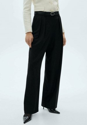 Kobieta ubrana w czarne, wysokie spodnie z szerokimi nogawkami i plisami, czarny pasek, kremową kurtkę na guziki oraz czarne szpilki z ostrym noskiem.