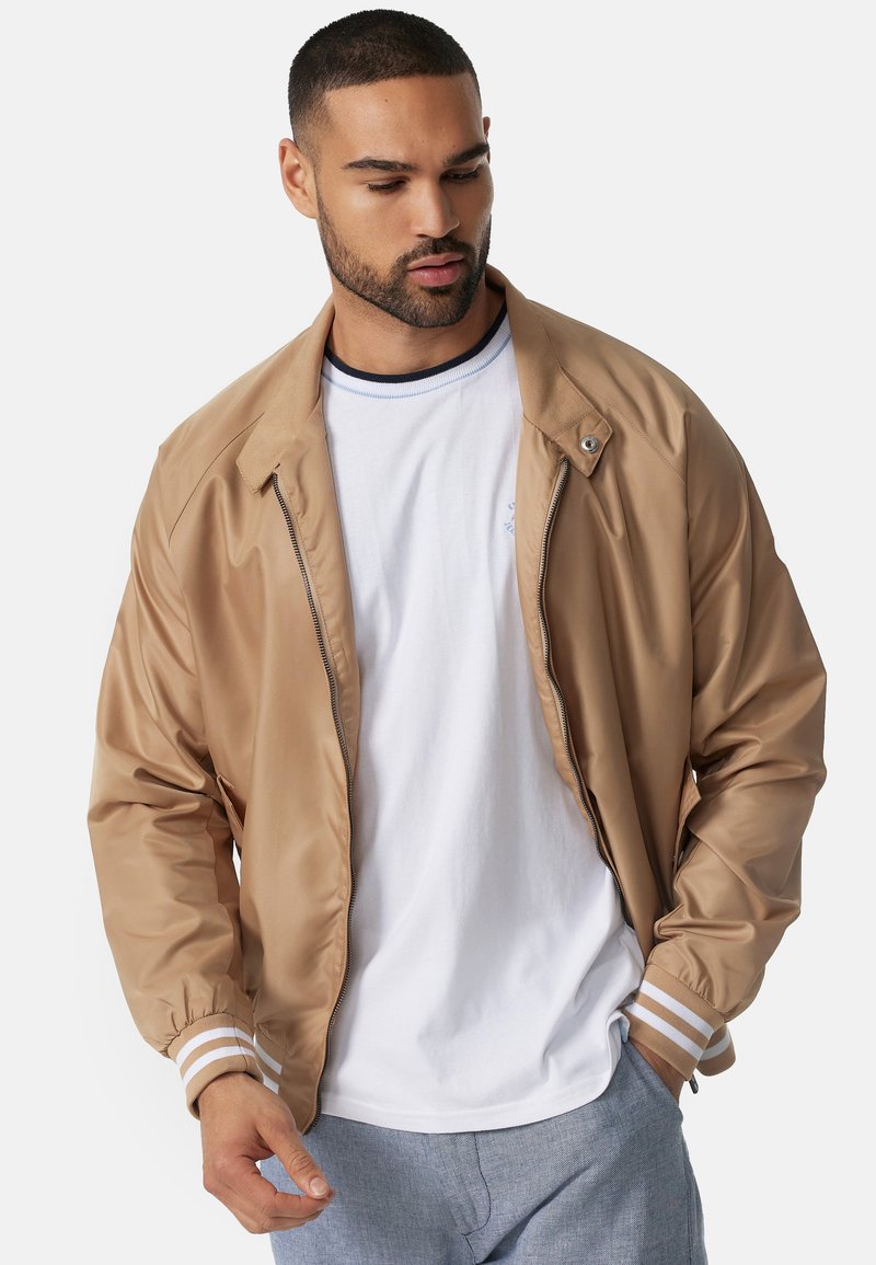 Cazadora bomber marrón con un acabado suave y brillante, con puños a rayas y cierre de cremallera, encima de una camiseta blanca.