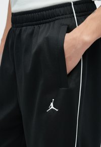 Čierne športové nohavice s elastickým pásom, bočnými vreckami, bielym lemovaním a bielym logom Jumpman na ľavom stehne. Hladká textúra.