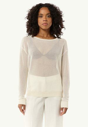 Lightweight cream sweater with open knit texture, ribbed cuffs, and a relaxed fit. Worn over a dark tank top and styled with beige trousers.