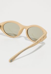 Ochelari de soare beige cu formă de ochi de pisică, lentile verzi, finisaj mat neted și braț curbat cu detalii decupate pe lungime.
