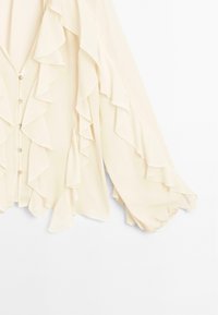 Blouse crème avec des volants en cascade sur le devant et les manches, fermeture boutonnée à l'avant, et poignets larges froncés.