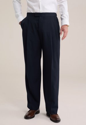 Homme portant un pantalon habillé noir plissé, des chaussures en cuir marron et une chemise blanche boutonnée, une main dans la poche.