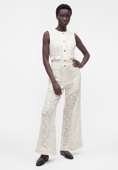Femme noire debout, main sur la hanche, portant un haut sans manches en dentelle blanche boutonné et un pantalon large assorti en dentelle avec des chaussures plates noires.