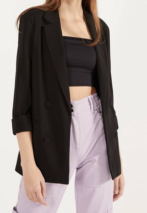 Femme portant un haut court tube noir, un blazer noir à double boutonnage avec les manches retroussées, et un pantalon taille haute violet clair.