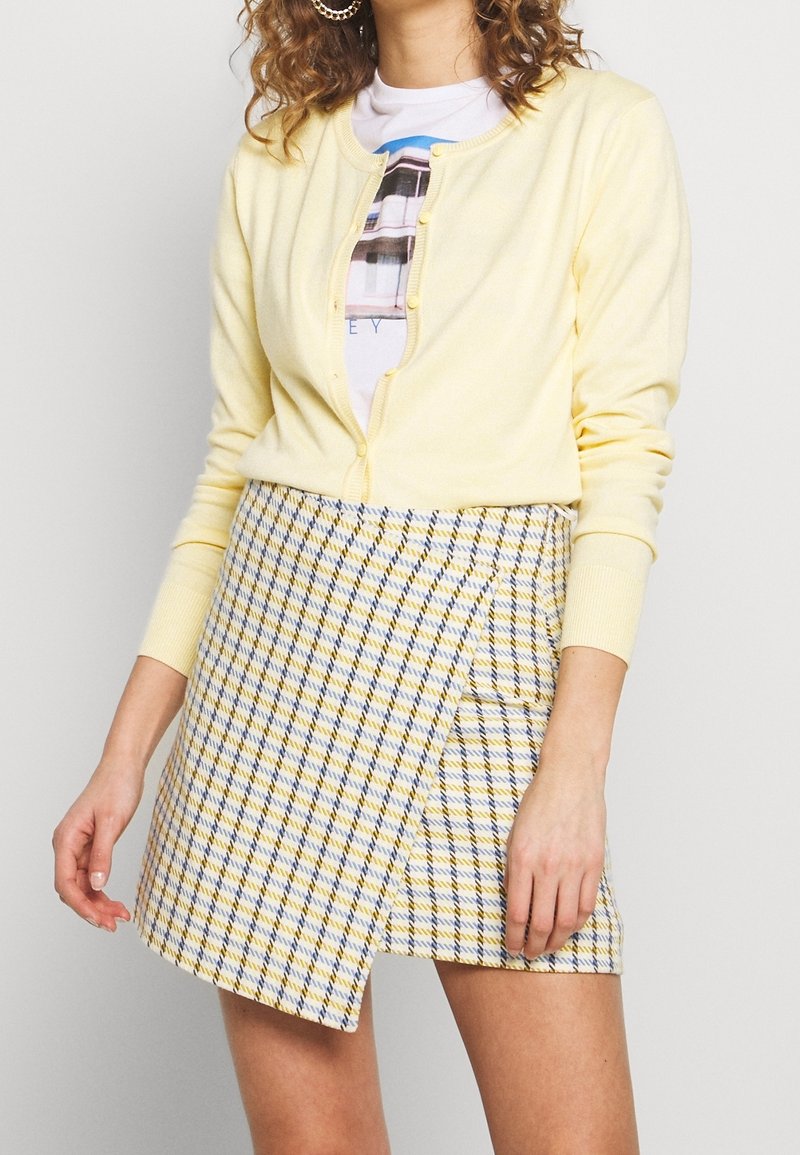 Jasnożółty sweter kardigan z okrągłym dekoltem i zapięciem na guziki, noszony z minispódniczką w kratę w odcieniach żółtego, niebieskiego i białego.