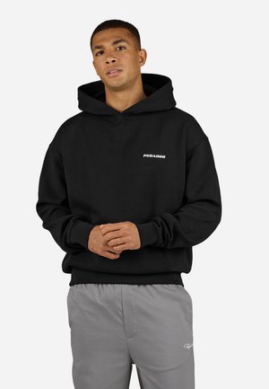 Man standing with hands clasped, wearing a black hooded sweatshirt with "PEGADOR" text and light gray pants against a plain background.