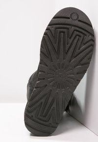 Semelle de botte noire avec des motifs de bande de roulement en zigzag et circulaires profonds reposant contre une surface d'angle blanche.