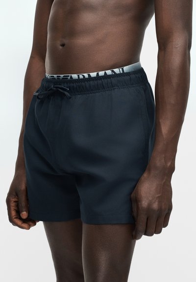 Άνδρας με σκουρόχρωμο δέρμα, με κορμό που φοράει μαύρο μαγιό σορτς με κορδόνι και εμφανή γκρι ζώνη εσωρούχου Armani, σε απλό φόντο.