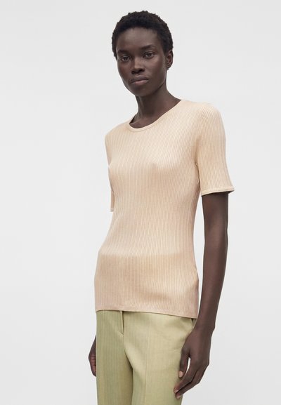 Γυναίκα με μπεζ ριπ κοντομάνικη μπλούζα και ανοιχτό ελιά παντελόνι, στέκεται μπροστά σε απλό λευκό φόντο.