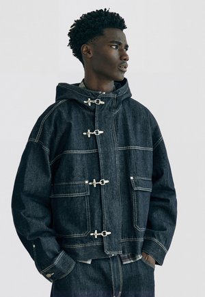 Giovane uomo con giacca di jeans scura con chiusure a bottoni e grandi tasche frontali, che guarda verso destra su uno sfondo semplice.