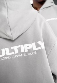 Felpa grigia con una texture morbida, caratterizzata da un grande testo bianco che recita "MULTIPLY APPAREL CLUB" sul retro. Dettaglio con zip visibile.