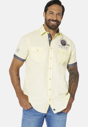 Man wearing a light yellow short-sleeve button-up shirt with pockets and blue jeans, smiling with one hand in pocket.