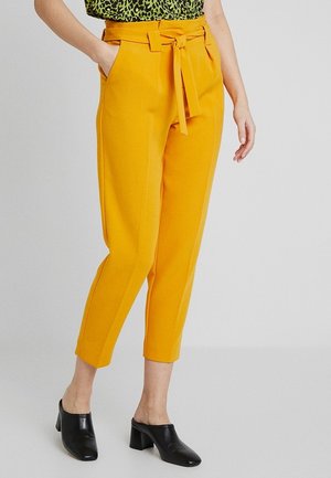 Żółte, dopasowane spodnie z wysokim stanem, paskiem do zawiązywania i bocznymi kieszeniami. Krótszy fason, gładka tekstura i czyste linie. Noszone są czarne mulesy.