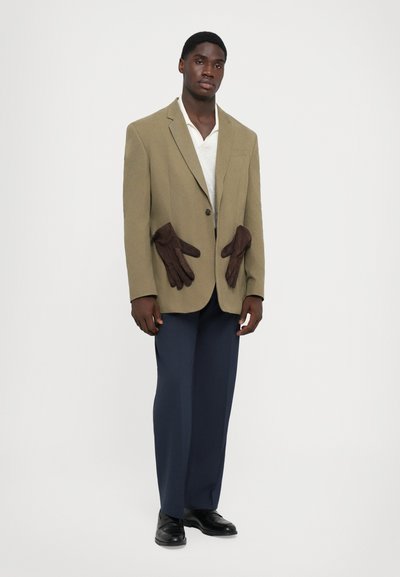 Beiger Blazer mit braunen Lederakzenten an den Taschen, kombiniert mit einem weißen T-Shirt und dunkelblauen Hosen. Schwarze Schuhe runden den Look ab.