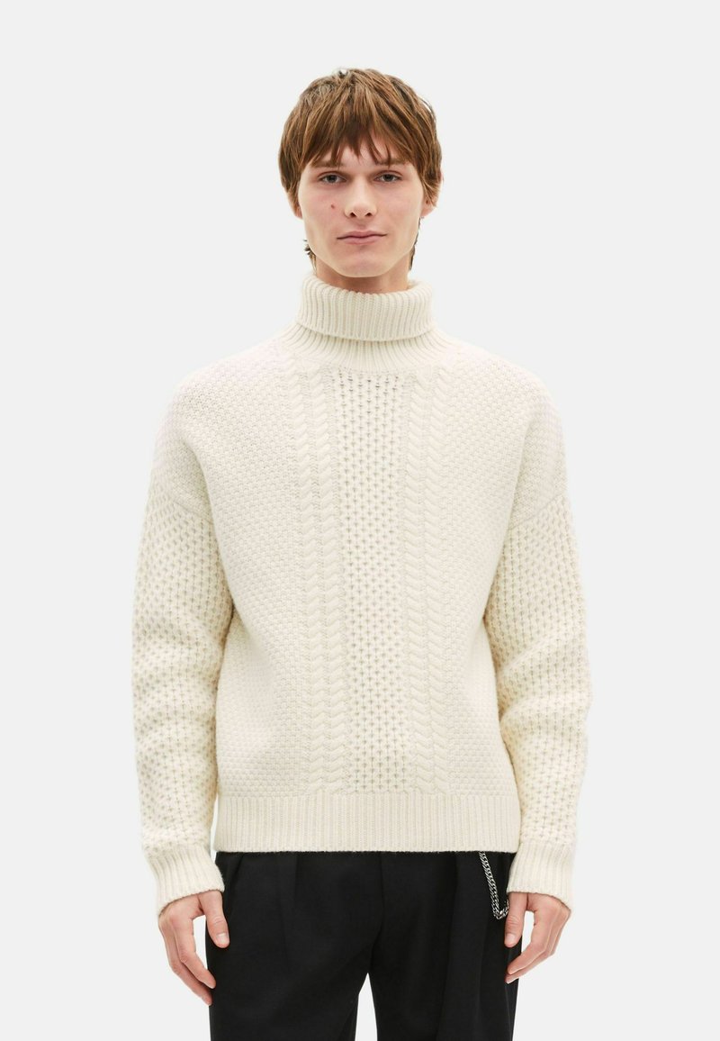 Pull en maille écrue, avec un col roulé haut, un motif torsad é au centre et des manches texturées. Poignets et ourlet côtelés.