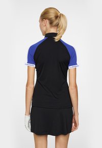 Svart sportsoverdel med blå och vita ärmar, med hög krage. Texten "J.LINDEBERG" på ärmen, i kombination med en svart kjol och en vit handske.