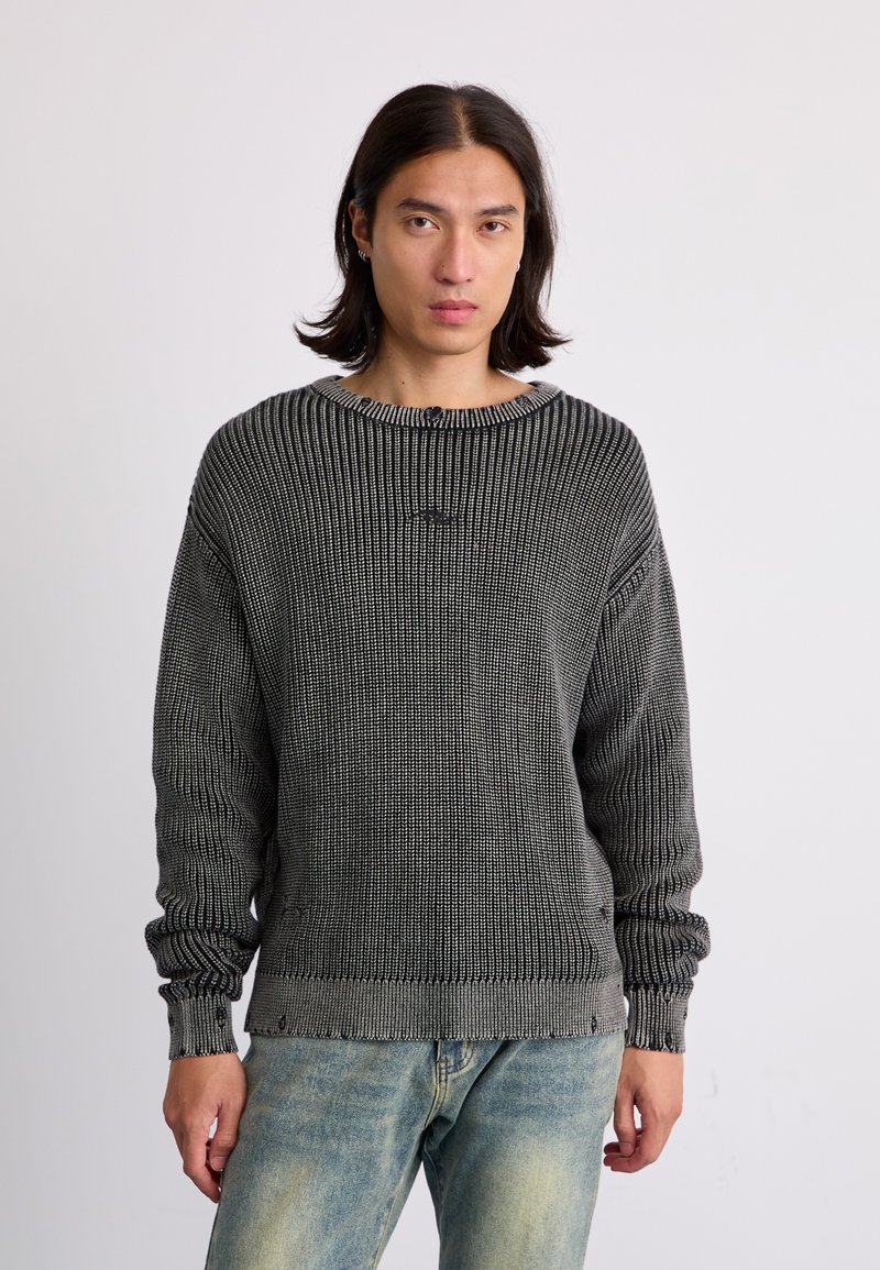 Pletena pulover v temno sivi barvi z teksturiranim vzorcem, širokim ovratnikom in rebrastimi manšetami, v kombinaciji z svetlimi jeans hlačami.