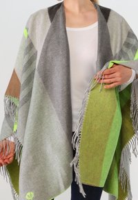 Personne portant un châle à franges multicolore avec des nuances de gris, vert, marron et jaune néon par-dessus une chemise blanche et un pantalon foncé.